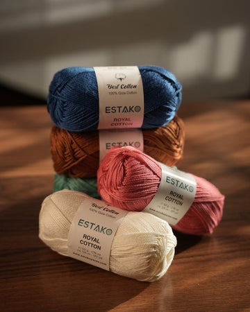 Estako Royal Cotton 50 g skeins stacked in blue, chocolate brown, rose and cream, with labels showing the 137 yard put-up
