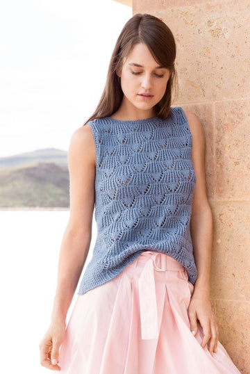 A model leaning against a sunlit stone wall by the sea, wearing a light blue openwork cotton summer top, a lightweight breathable knit.