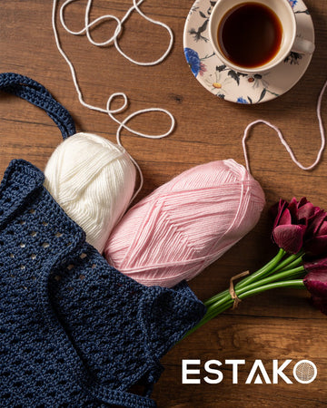 A navy crochet net bag with an open mesh stitch, beside cream and pink cotton skeins, a coffee cup and burgundy tulips on a wood table.